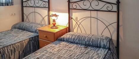 A room with two single beds, a wooden nightstand with a lamp, and a window with curtains.