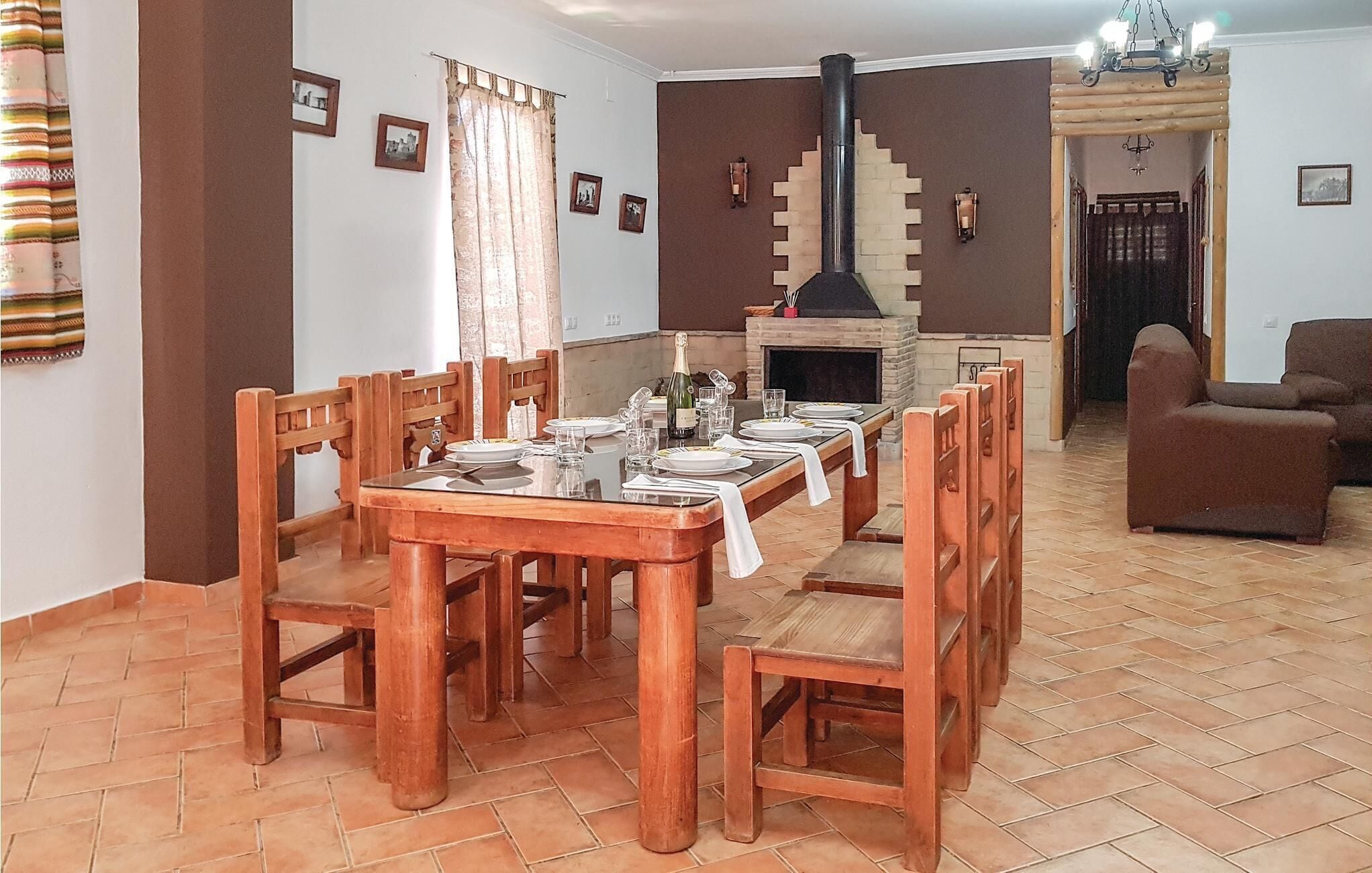 A dining area with a long wooden table set for six, surrounded by wooden chairs. A fireplace with a chimney is visible in the background.