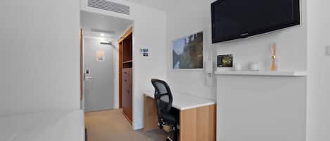 A compact hotel room with a single bed, a desk, a chair, a flat-screen TV, and a small shelf with decorative items.