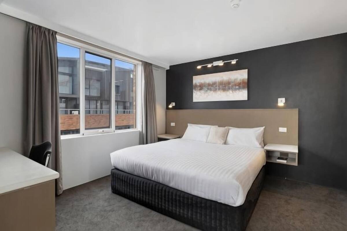 A hotel room with a large bed, a desk, a chair, a window with curtains, and a painting on the wall.