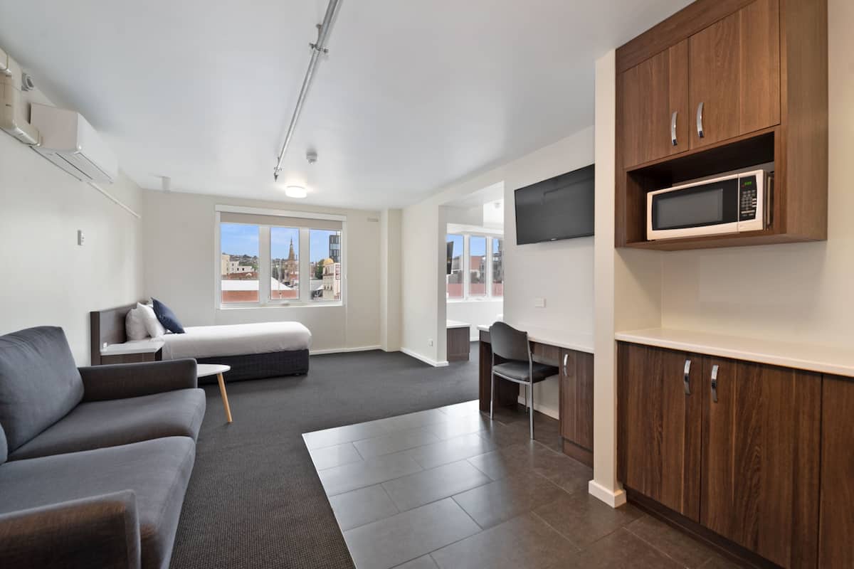 A modern living room with a sofa, a small table, a TV mounted on the wall, and a kitchen area with wooden cabinets and a microwave.