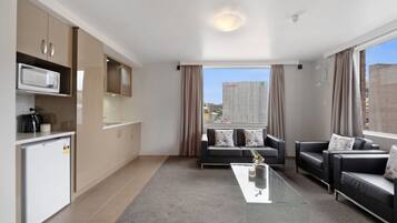 A modern living room with a glass coffee table, two sofas, a kitchen area with a microwave, and a view of a cityscape through large windows.
