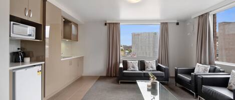 A modern living room with a glass coffee table, two sofas, a kitchen area with a microwave, and a view of a cityscape through large windows.
