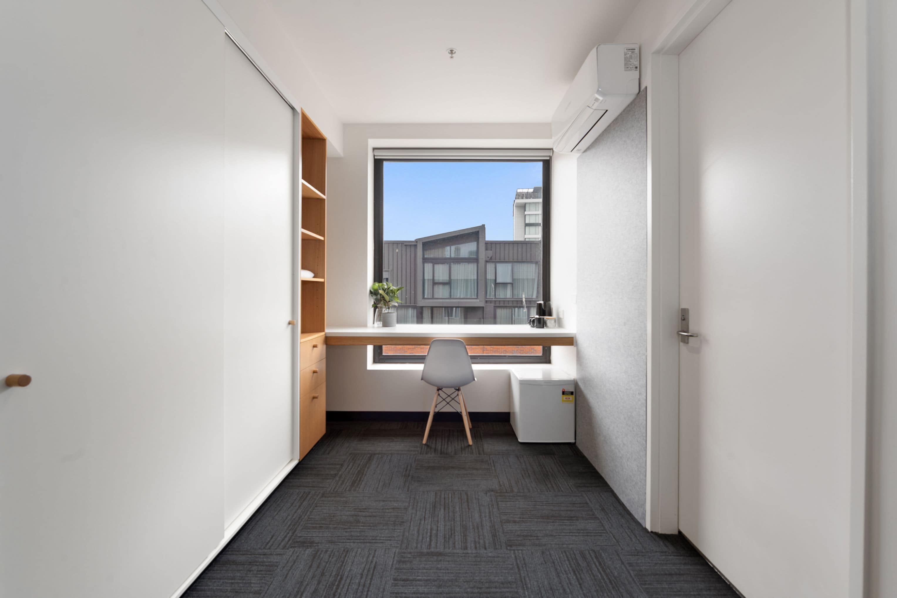 Een kleine kamer met een bureau, stoel en een raam met uitzicht op een gebouw.