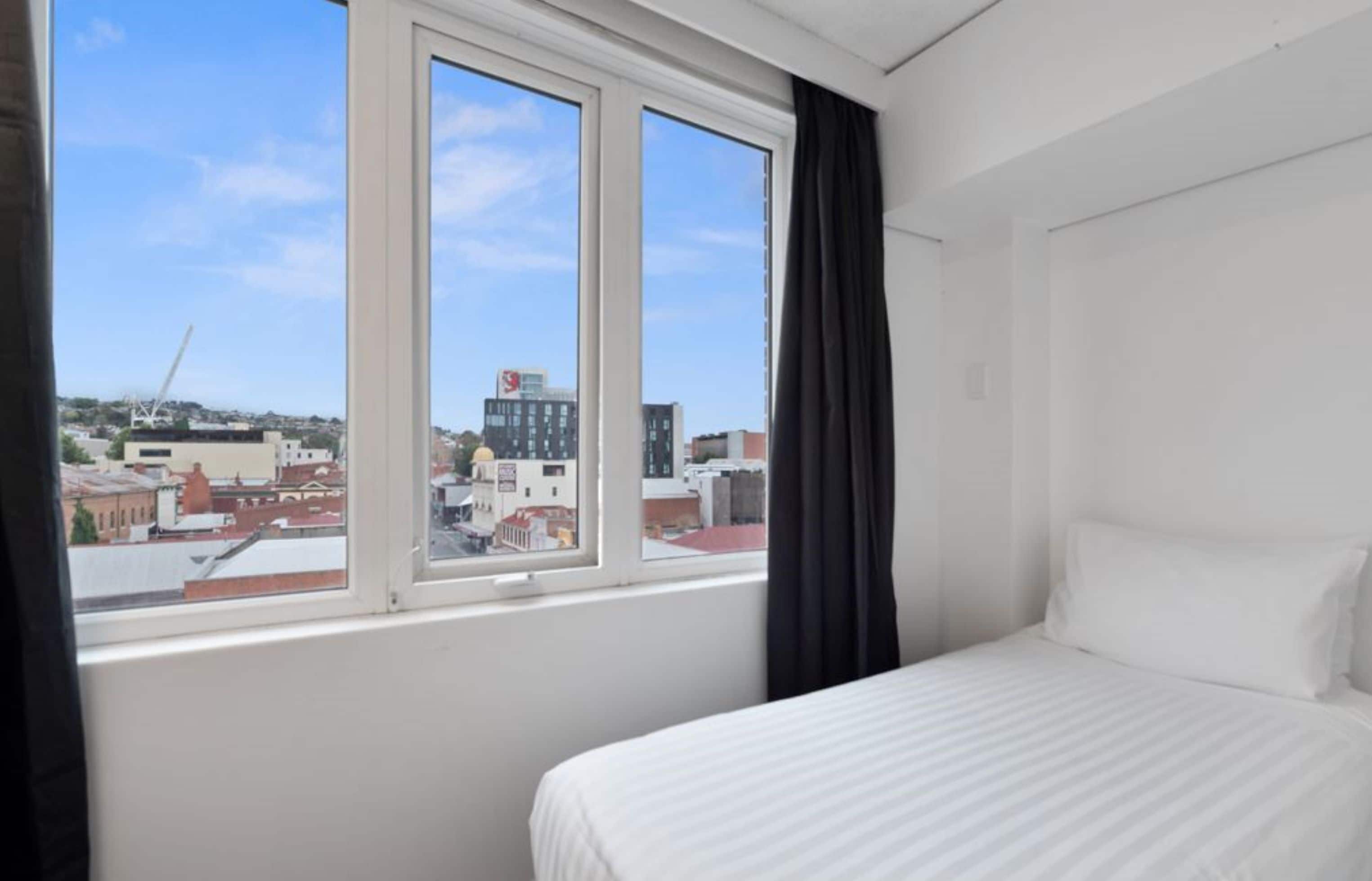 A hotel room with a large window, a bed, and a view of the cityscape.