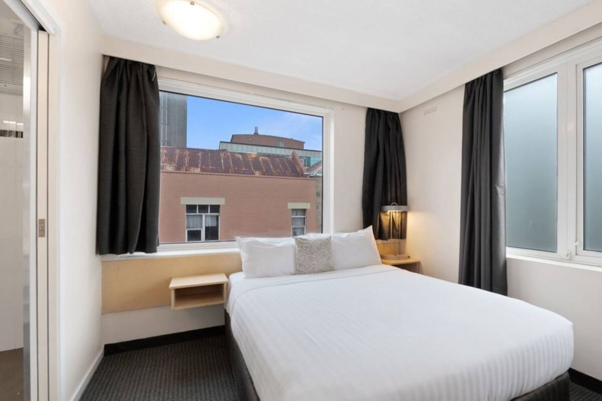 A hotel room with a large bed, a bedside table with a lamp, a window with curtains, and a view of a building outside.