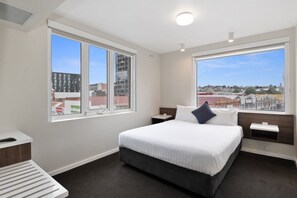 A modern hotel room with a large bed, two bedside tables, a nightstand, a window with a city view, and a small desk with a chair. - Allurity Hotel (Hobart)