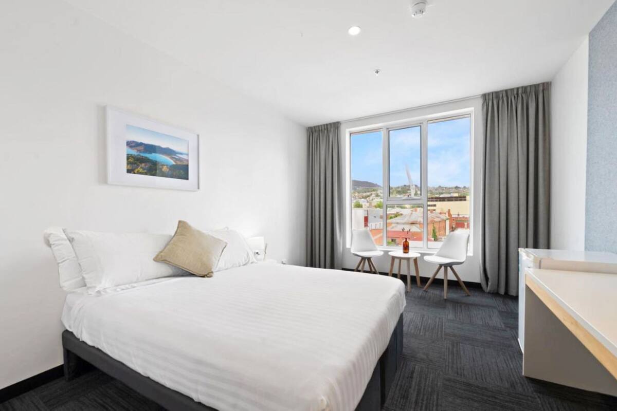 A hotel room with a bed, a desk, two chairs, a small table, a window with a view, and a framed picture on the wall.