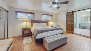 Un dormitorio con cama, dos lámparas de noche, ventilador de techo, puerta corredera estilo granero y un baño visible a través de la puerta.