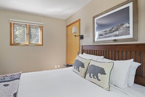 Een slaapkamer met een bed, twee kussens met berenmotieven, een nachtkastje met een lamp, een ingelijste foto aan de muur en een raam met uitzicht op bomen.