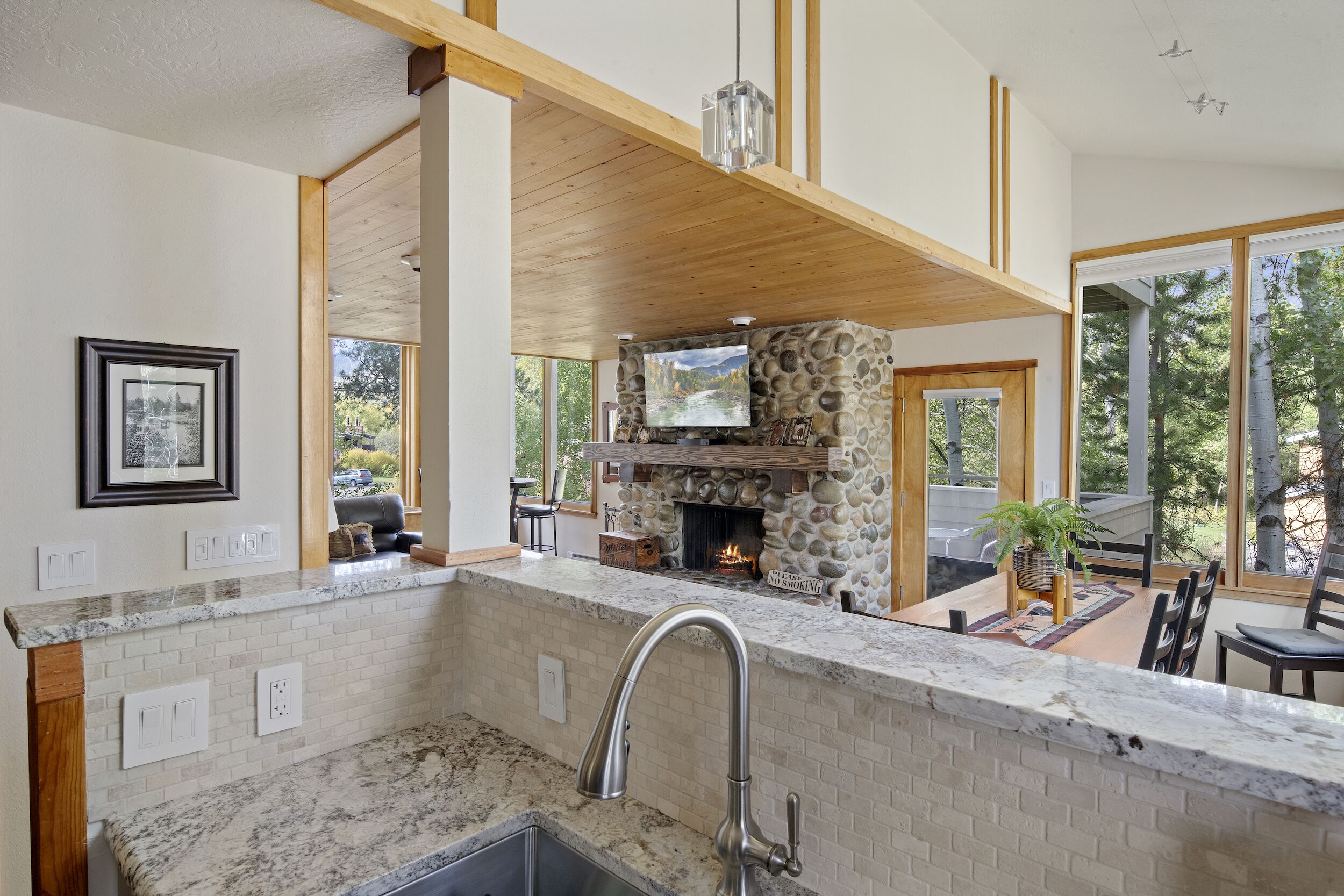 A kitchen with a stone fireplace, wooden beams, and a large window overlooking a deck.