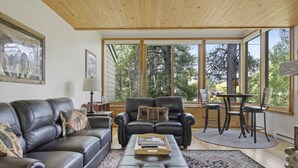 Un salón con un sofá de cuero, un sillón a juego, una mesa de centro y un comedor con una mesa y sillas. Amplias ventanas ofrecen vistas al exterior.