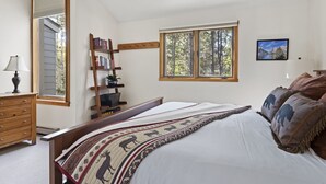 Un dormitorio con una cama grande, una escalera de madera, una estantería, una ventana con vista a árboles y un cuadro en la pared.