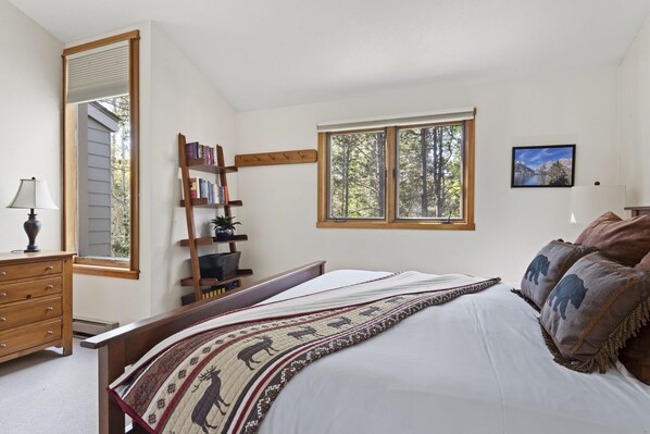 Un dormitorio con una cama grande, una escalera de madera, una estantería, una ventana con vista a árboles y un cuadro en la pared.
