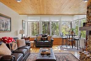 A spacious living room with a wooden ceiling, large windows, a black leather sofa, a coffee table, and a stone fireplace. - Outpost: Yampa 2424 - Aspens Location! (Wilson)
