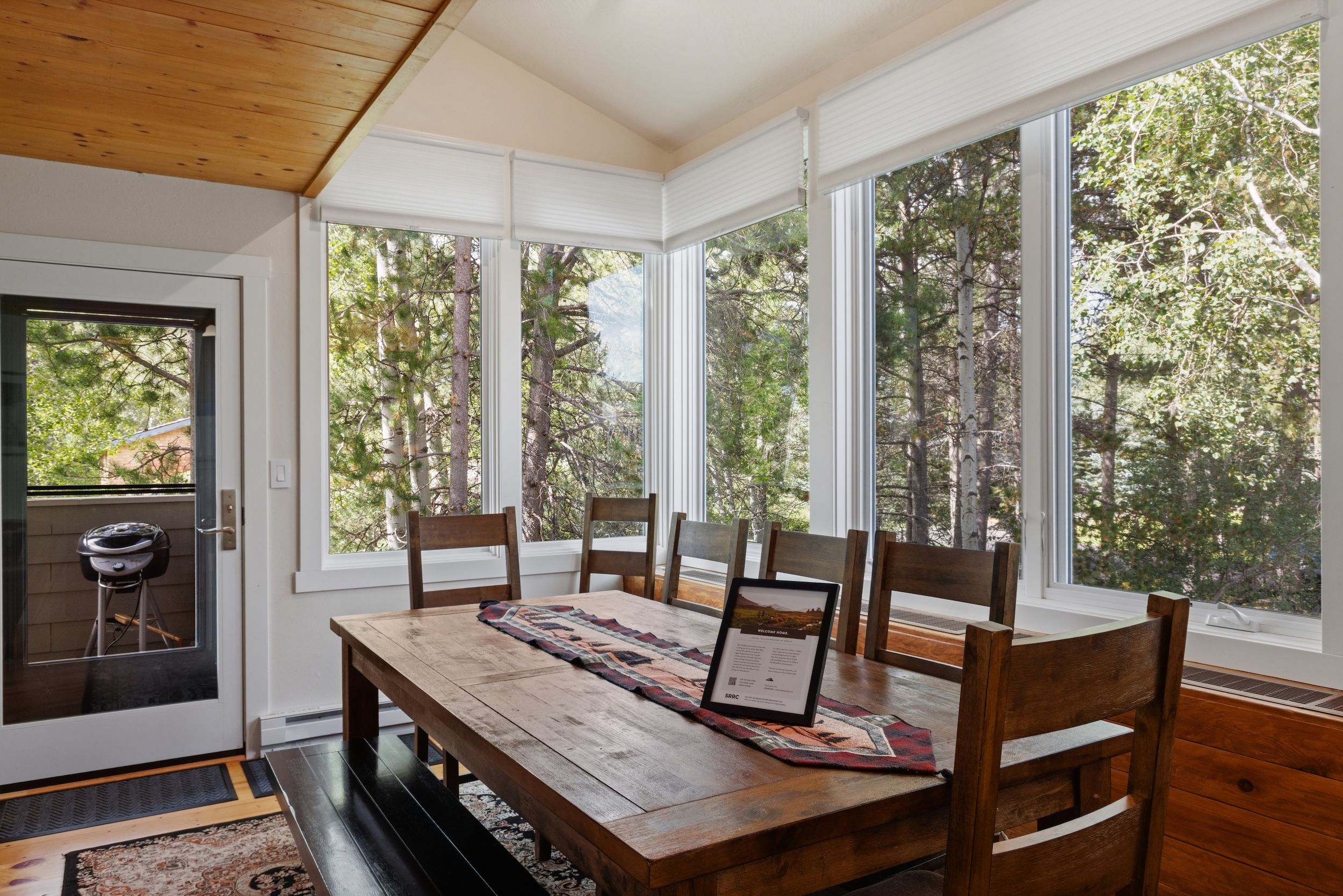 A dining area with a wooden table and chairs, large windows, and a view of trees.