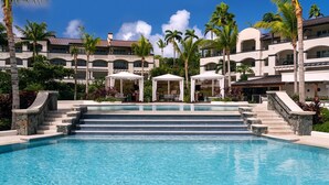 Piscina de resort con agua azul cristalina, rodeada de palmeras y un edificio de varios pisos con balcones.