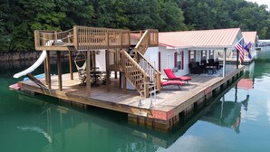Un muelle de madera con un tobogán, mobiliario exterior y un área cubierta con banderas.