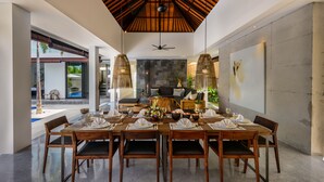 A dining area with a long wooden table set for a meal, a ceiling fan, and a stone wall in the background. - Villa Waha (Canggu)