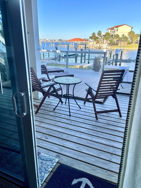 A wooden deck with outdoor seating, including two lounge chairs and a small table, overlooking a marina with boats and a building in the background.