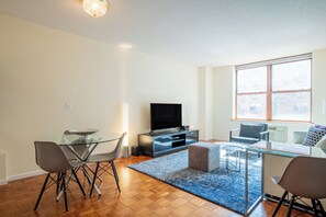 A modern living room with a glass dining table, comfortable seating, a flat-screen TV, and large windows. - One Bedroom in Waterfront Building with NYC Views (Hoboken)