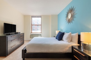 A bedroom with a bed, a TV on a dresser, a window, and a wall clock. - One Bedroom in Waterfront Building with NYC Views (Hoboken)