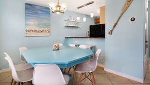 A dining area with a blue table and white chairs, a beach painting, a wooden paddle on the wall, and a kitchen in the background. - Emerald Shores: Steps from the Gulf, Navarre Beach (Navarre)