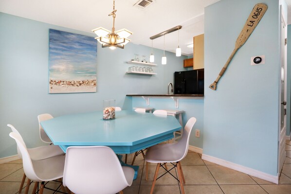 A dining area with a blue table and white chairs, a beach painting, a wooden paddle on the wall, and a kitchen in the background.