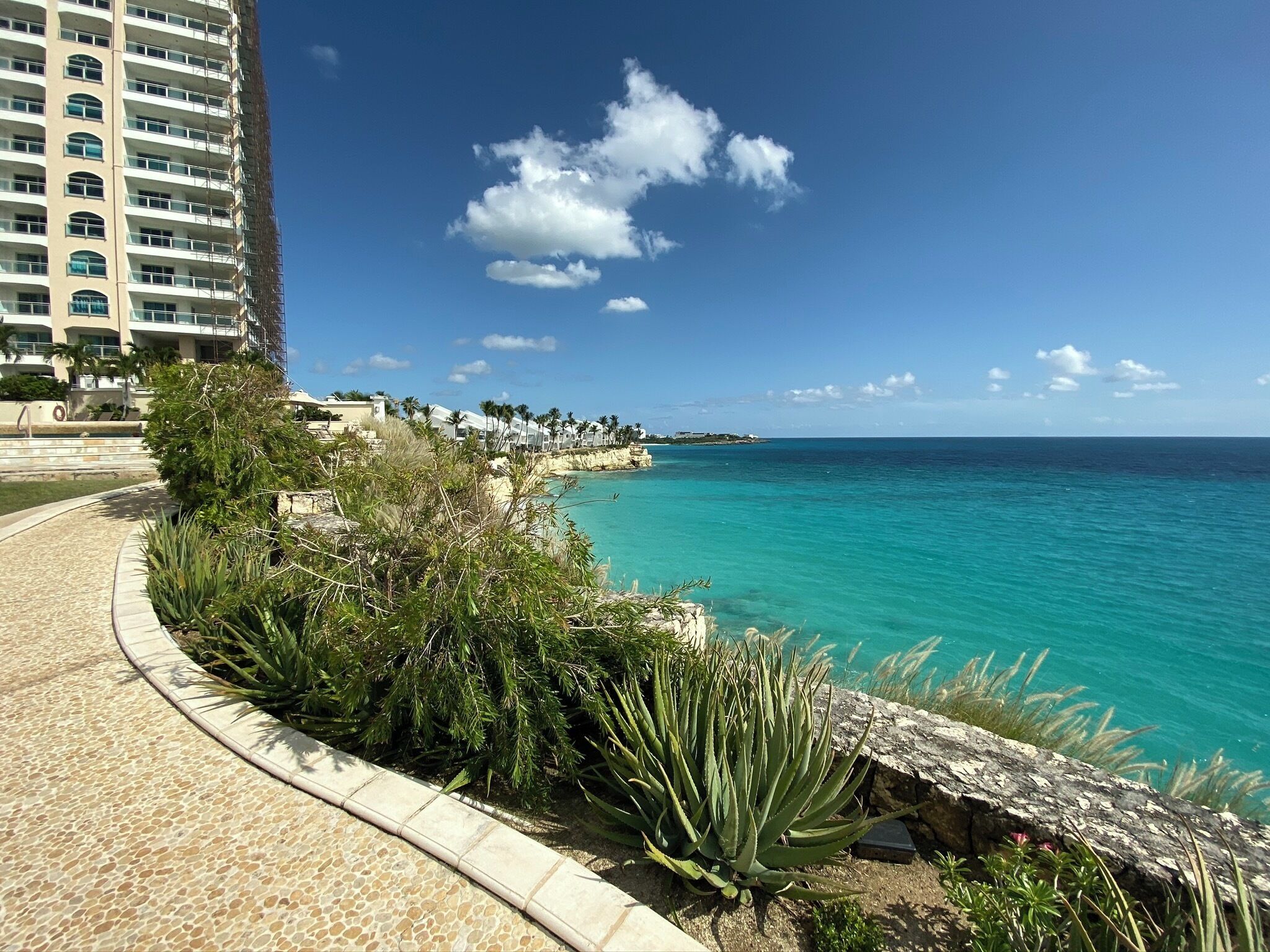A coastal walkway with a clear view of the ocean, a building with balconies, and a rocky shoreline.