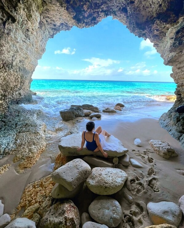A person sitting on rocks at a beach with a clear view of the ocean through a cave-like opening. - Ocean Bliss with Balcony and Peloton by Premium at Rainbow Beach Club (Cupecoy)