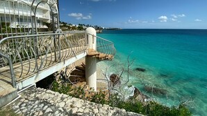 Un hotel frente al mar con balcón que da a aguas turquesas, una escalera hacia la playa y vistas despejadas del océano.
