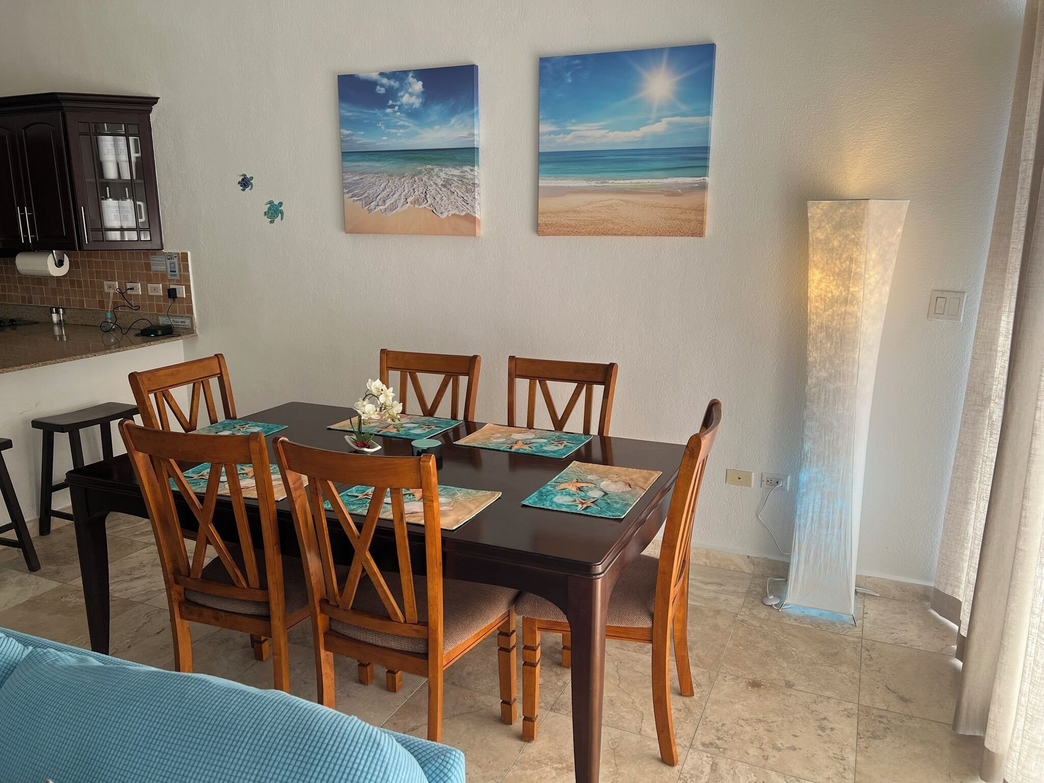 A dining area with a wooden table and chairs, a kitchen in the background, and two beach-themed wall art pieces.