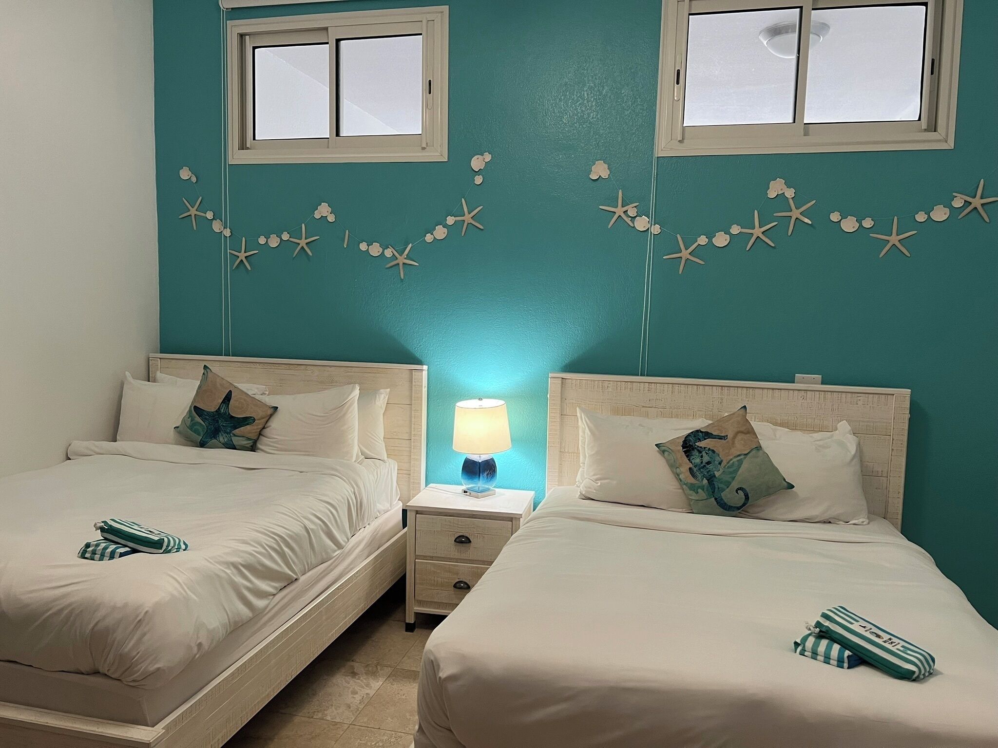 A hotel room with two beds, a nightstand with a lamp, and a wall decorated with starfish and cloud motifs.