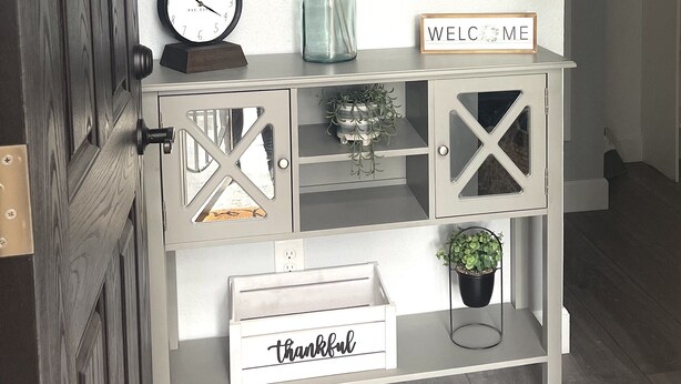 Una consola blanca con un reloj, un letrero que dice 'WELCOME', un letrero que dice 'THANKFUL', y objetos decorativos como un florero y una planta.