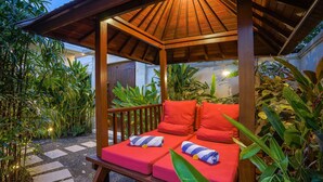 A wooden gazebo with a thatched roof, two red cushioned benches with striped towels, surrounded by lush greenery and a stone pathway.