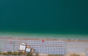 Une vue aérienne d’une plage avec des rangées de parasols blancs et quelques chaises de plage, un petit bâtiment et une chaise rouge près de l’eau.