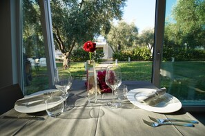 Une table à manger dressée avec des assiettes, des verres et des couverts, agrémentée d’un vase de fleurs et d’une rose, visible à travers une fenêtre donnant sur un jardin.