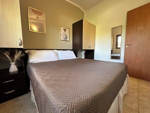 A neatly made bed with a patterned bedspread, a bedside table with a vase of dried flowers, and a wall-mounted artwork.