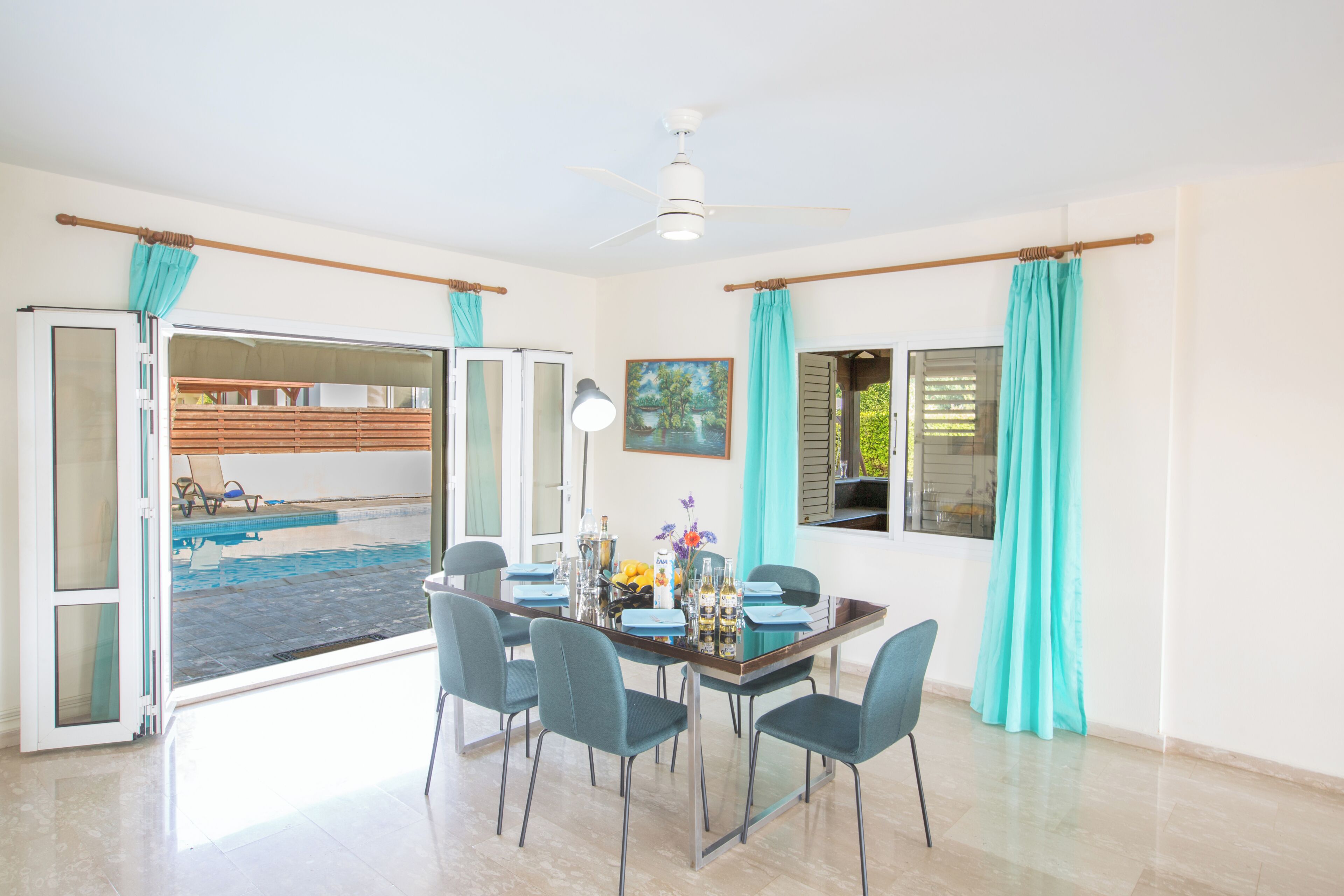 A dining area with a glass table, four chairs, a ceiling fan, and a view of a swimming pool.