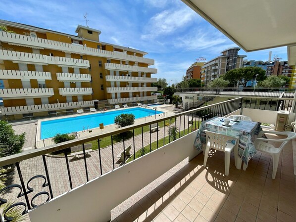 Un balcón con vista a una piscina, una mesa con sillas y un edificio con balcones al fondo.