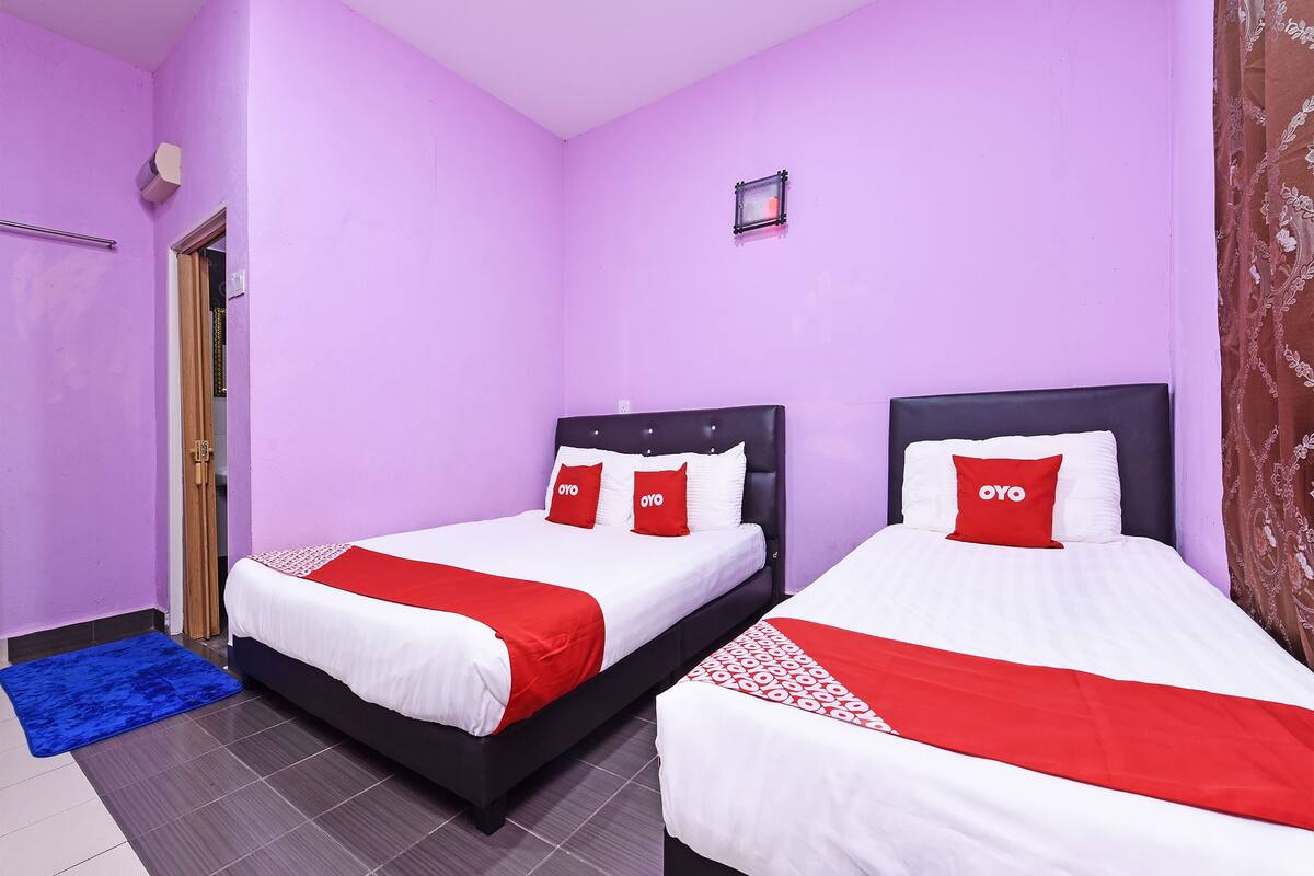 A room with two beds, each with a black headboard, white bedding, and red pillows. The walls are purple, and there is a small window with a picture frame.
