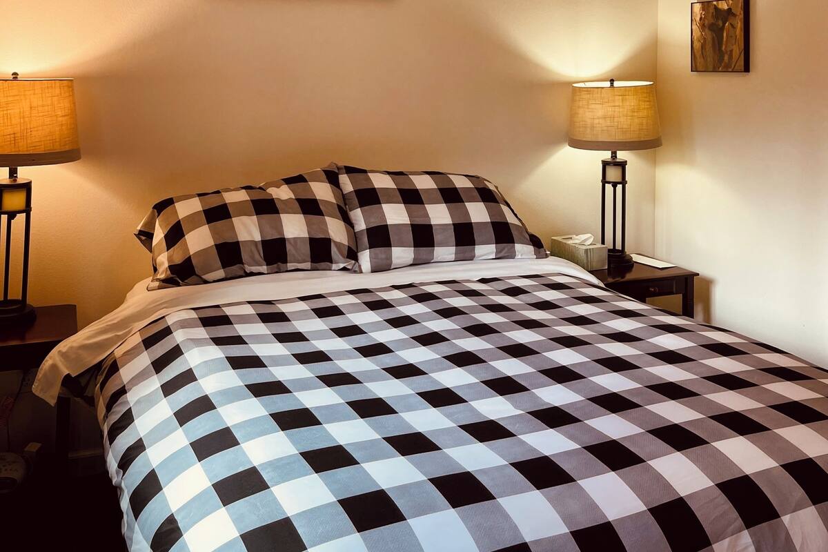 A hotel room with a checkered bedspread, two bedside lamps, a wall-mounted sign reading "Denali National Park," and a small framed picture on the wall.