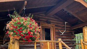 Una cabaña rústica de troncos con un porche de madera, una maceta colgante con flores y una escalera de madera.