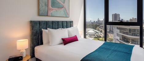 A modern hotel room with a large bed, a city view, a painting on the wall, and a small table with a lamp.