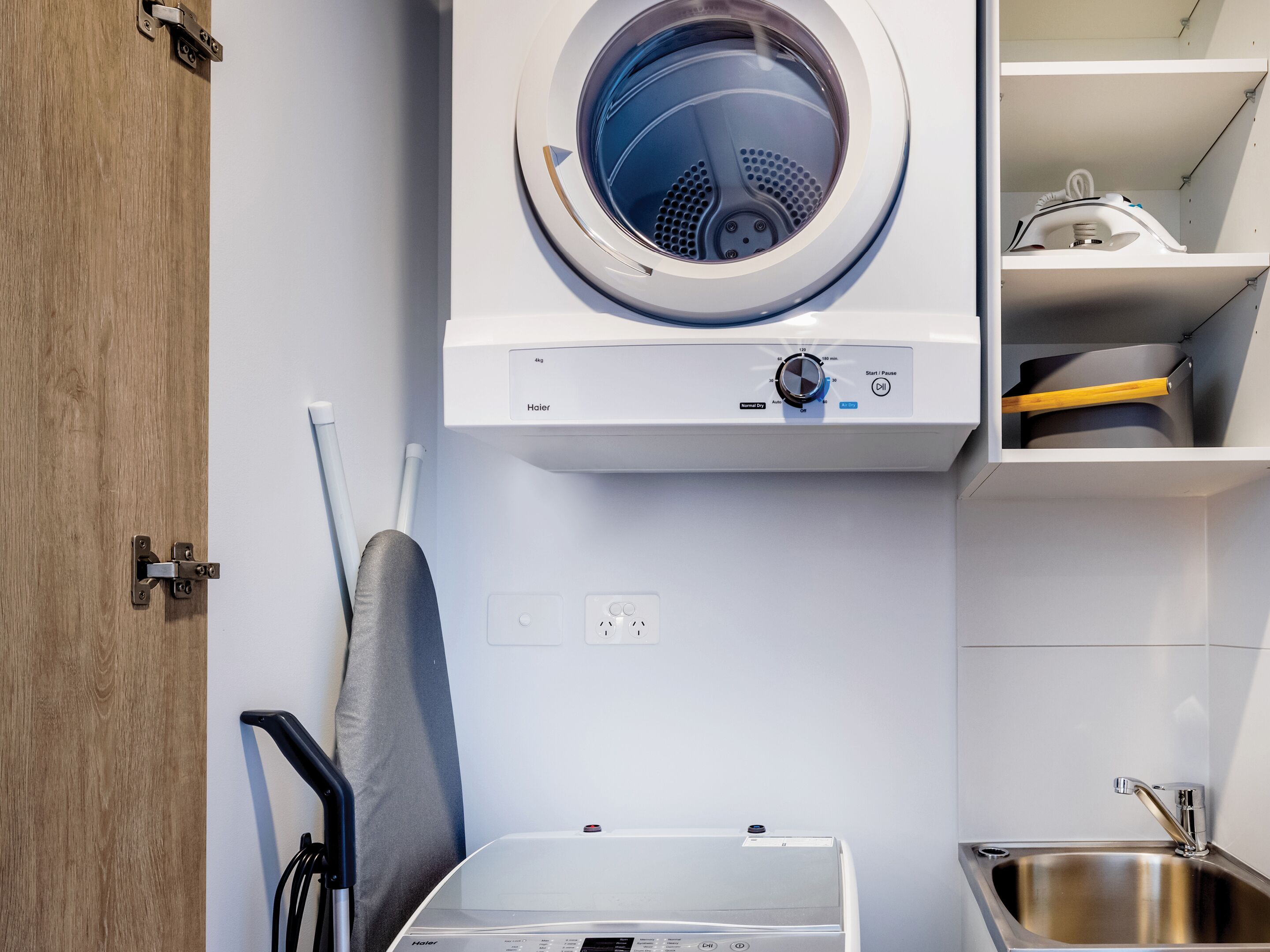 A compact laundry area with a front-loading washer and dryer, a small ironing board, and a sink.