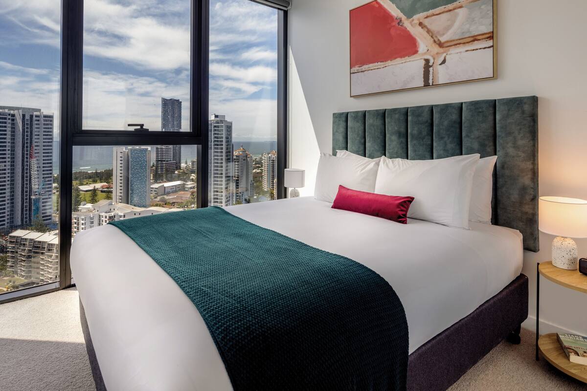 A hotel room with a large bed, a view of the cityscape through a floor-to-ceiling window, and a modern headboard.