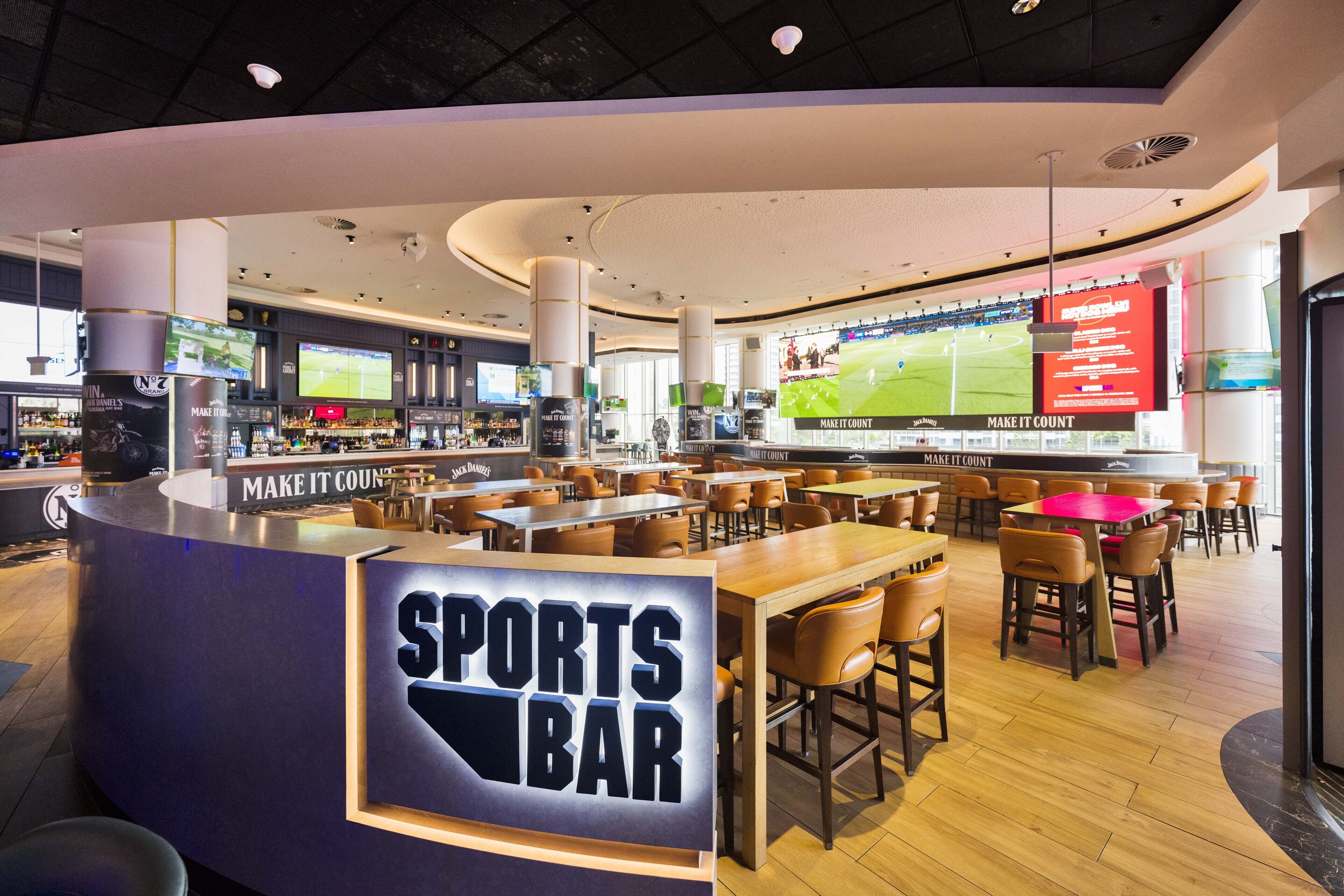 En sportbar med flera sittområden, en bardisk med en skylt som visar att det är en sportbar, och stora skärmar som visar sportevenemang.