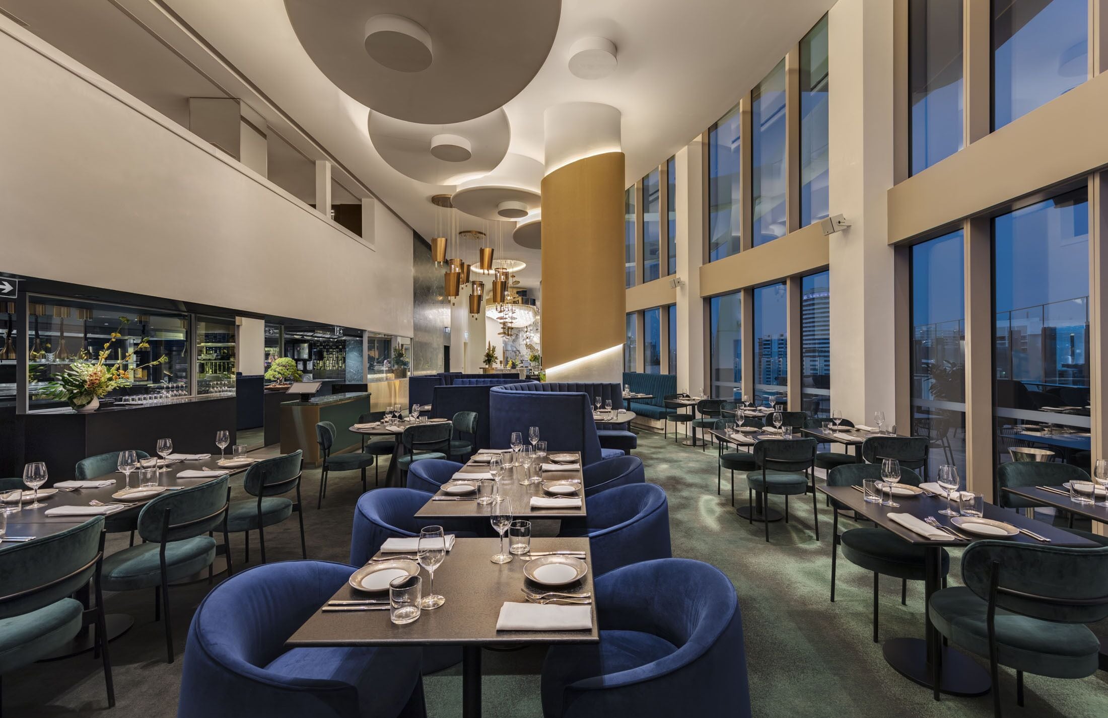 Un restaurante moderno con varias mesas y sillas, una zona de bar, y grandes ventanales que ofrecen vistas a la ciudad.