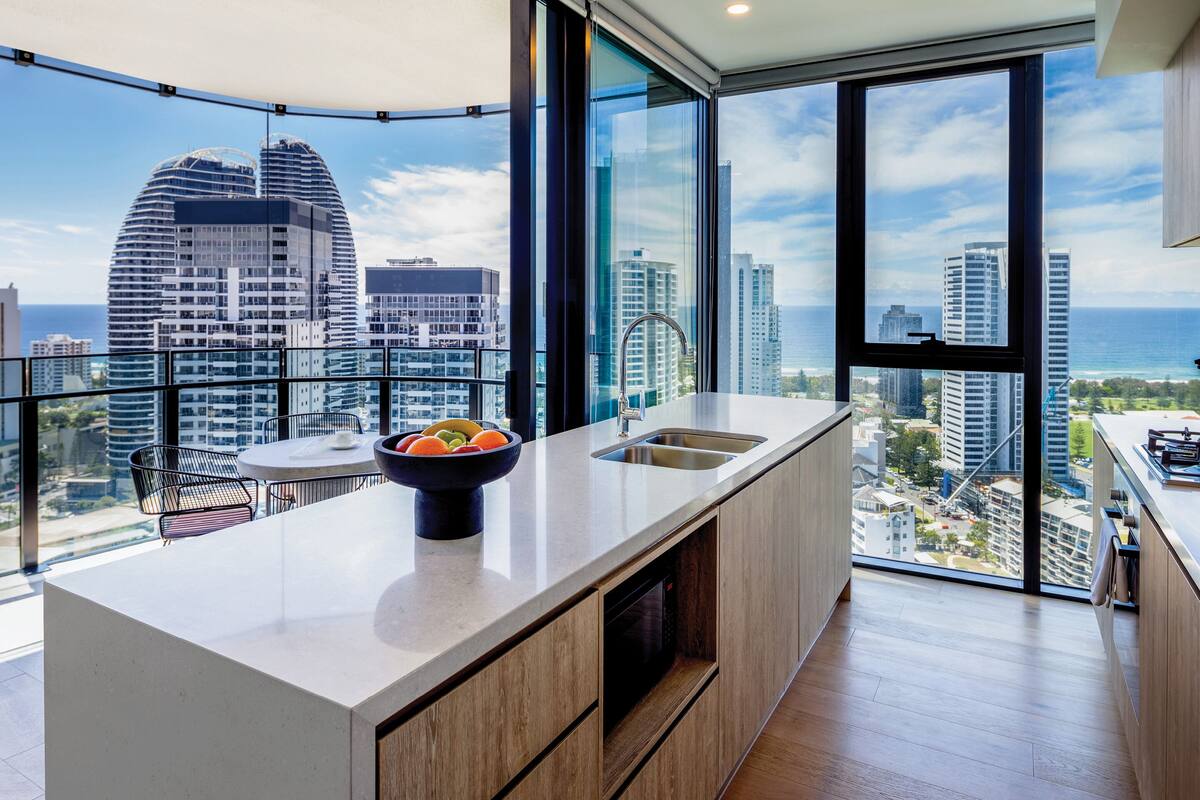 A modern kitchen with a large island, a bowl of fruit, and a city view through large windows.