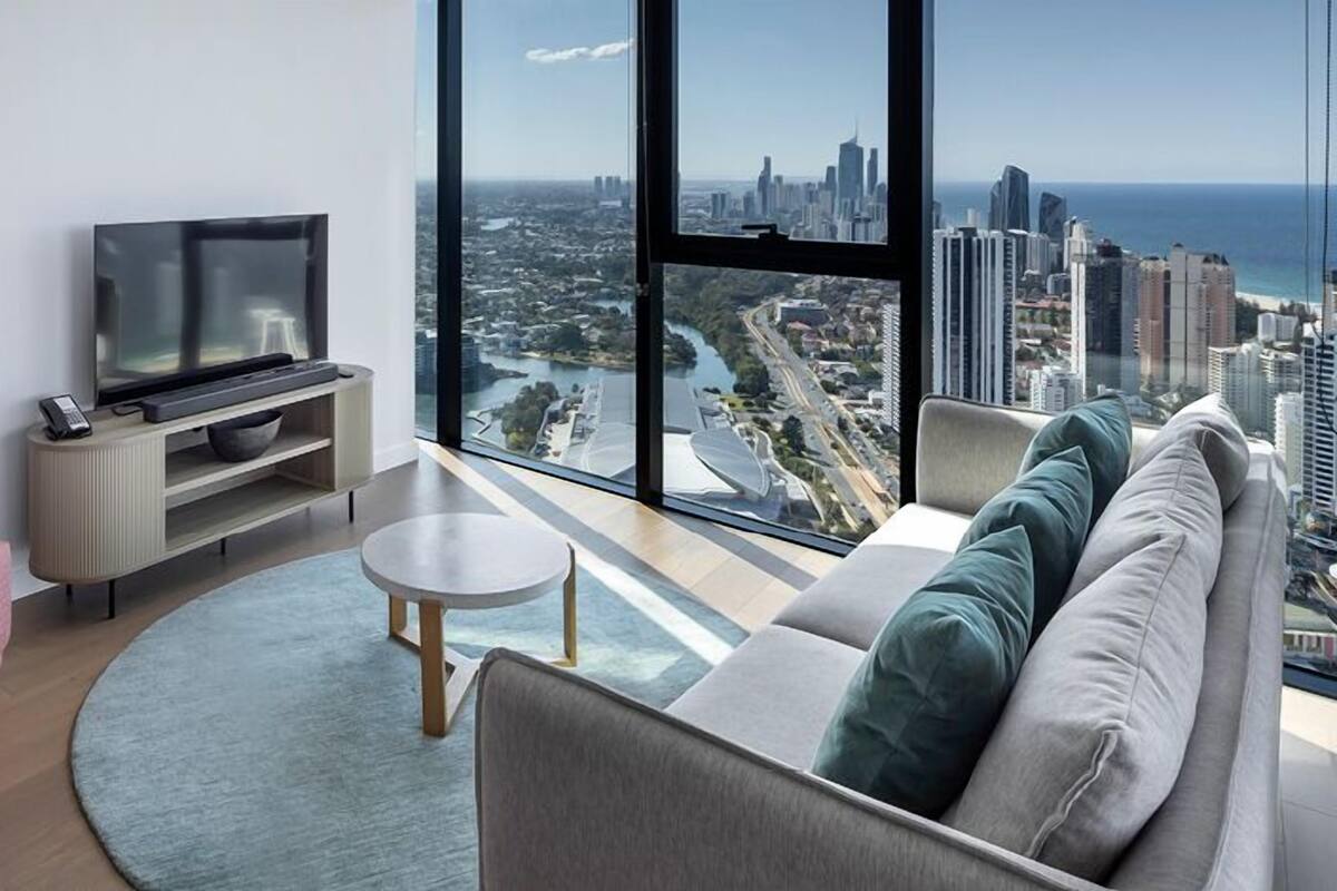 A modern living room with a pink armchair, a grey sofa, a flat-screen TV on a wooden stand, and a large window offering a city view.